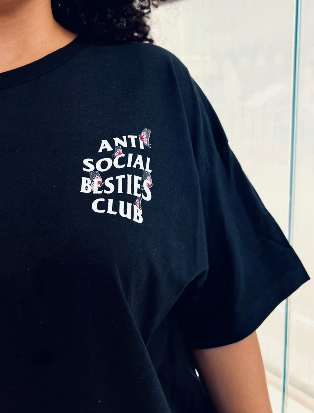 Front close-up of the black AntiSocial Besties Club Butterfly Tee by Dass My Best Friend. The minimalist design features the bold "Antisocial Besties Club" print adorned with small butterfly details, adding a playful and stylish touch. Perfect for introverted besties embracing their unique vibe. The soft black tee offers a casual yet statement-making look.