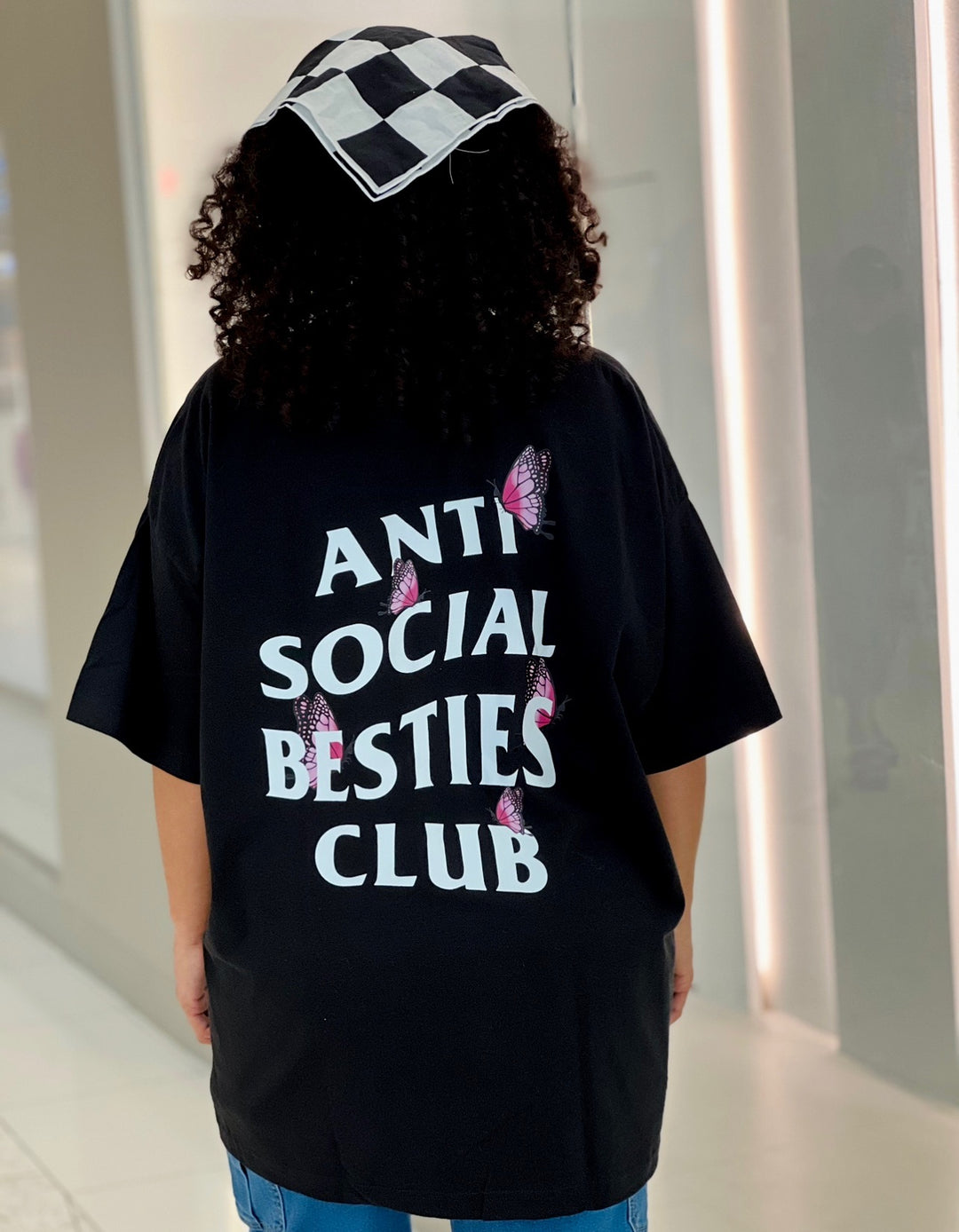 Woman standing in a mall wearing the AntiSocial Besties Club Butterfly Tee by Dass My Best Friend, showing the back design of the tee. The butterfly details add a playful twist to the iconic design, perfect for introverted best friends with an edgy style.