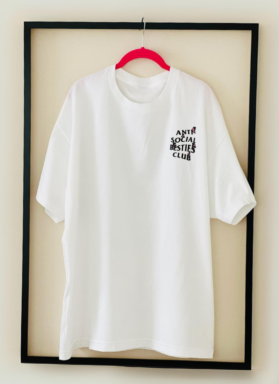 Front view of the white AntiSocial Besties Club Butterfly Tee by Dass My Best Friend, hanging inside a picture frame. The bold AntiSocial Besties Club design with butterfly details is displayed, offering a playful twist on the classic tee for best friends.