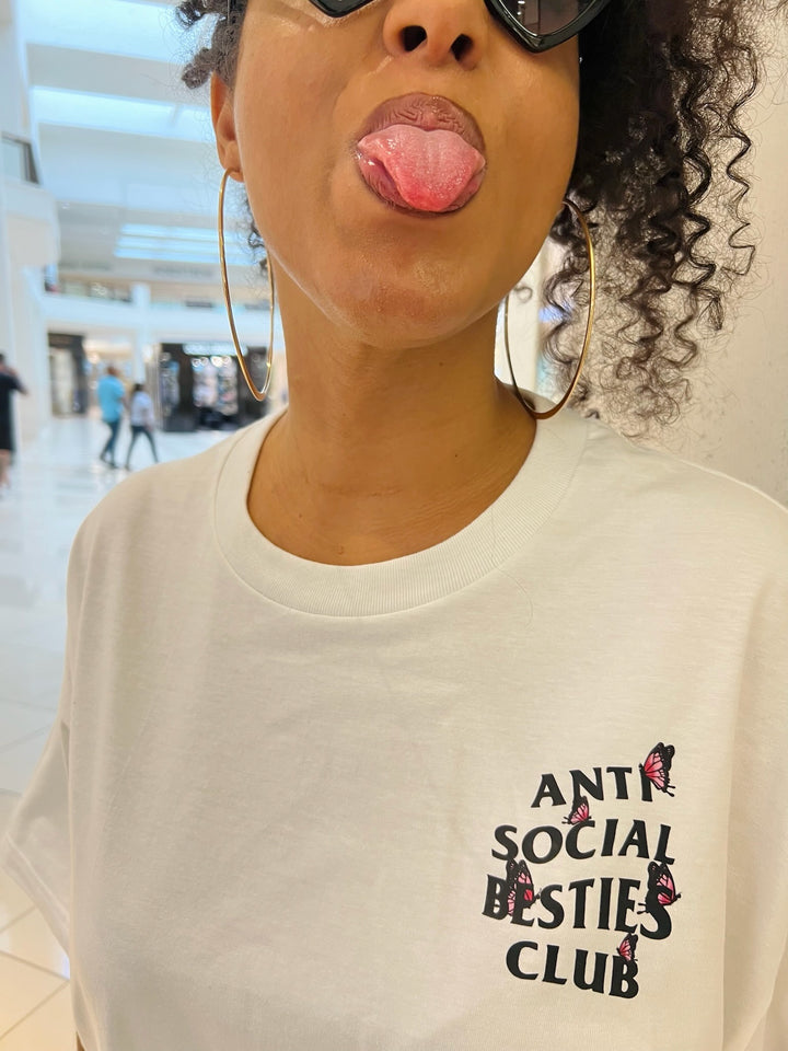 Front close-up of the white AntiSocial Besties Club Butterfly Tee by Dass My Best Friend. The model playfully sticks her tongue out, showcasing the butterfly design and bold "Antisocial Besties Club" print. A fun and edgy look for introverted best friends.