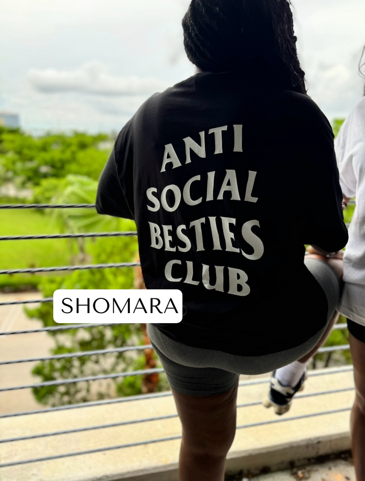 Shomara leaning on a fence, looking away from the camera, wearing the black AntiSocial Besties Club Tee by Dass My Best Friend. The bold "Antisocial Besties Club" print on her back stands out, capturing the edgy, introverted vibe perfect for best friends.