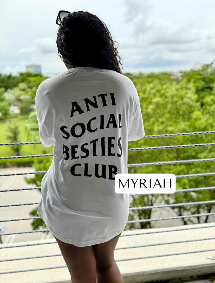 Myriah standing at a fence with her back facing the camera, wearing the white AntiSocial Besties Club Tee by Dass My Best Friend. The bold "Antisocial Besties Club" print on her back is clearly visible, showcasing the playful and introverted style designed for best friends.