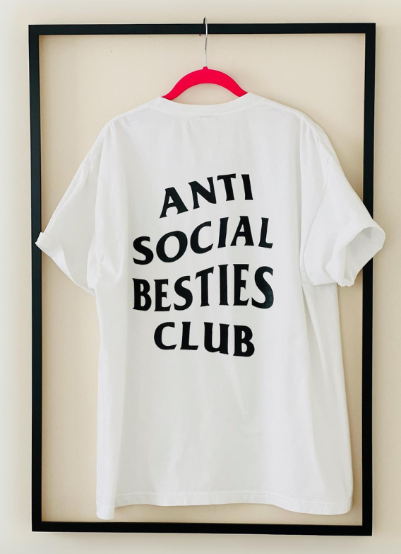  white AntiSocial Besties Club Tee by Dass My Best Friend. The bold "Antisocial Besties Club" print on her back is clearly visible, showcasing the playful and introverted style designed for best friends.