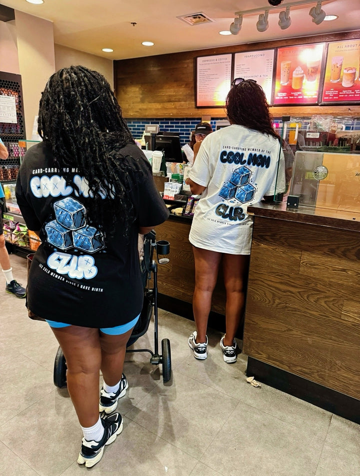 Two moms buying coffee at Starbucks, one wearing the black Cool Mom Club Tee and the other in white by Dass My Best Friend. The relaxed-fit tee features the bold "COOL MOM CLUB" graphic on the front and back, perfect for confident, laid-back moms.