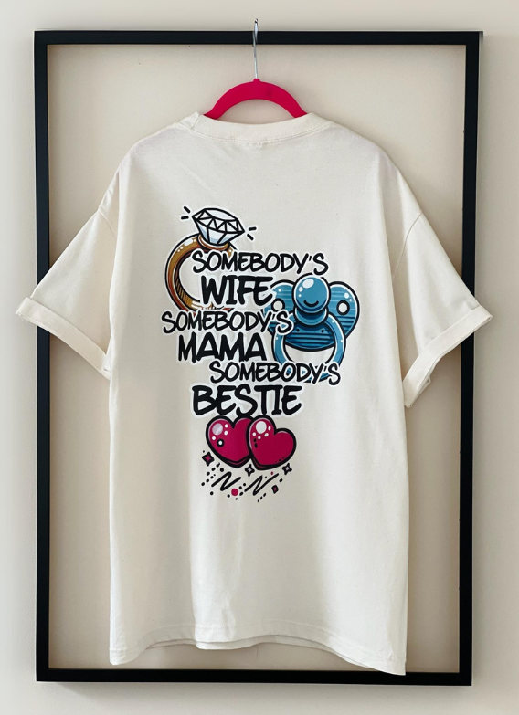 Rear view of the white "Somebody's Wife, Mama, Bestie" Tee by Dass My Best Friend, hanging inside a picture frame. The playful graphic is displayed on the back, celebrating the important roles of wife, mom, and best friend in a relaxed, stylish fit.