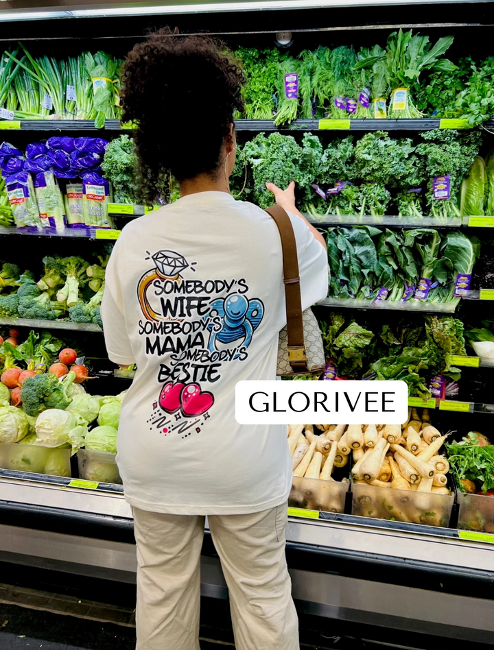 Back view of Glorivee wearing the white "Somebody's Wife, Mama, Bestie" Tee by Dass My Best Friend, shopping for vegetables in Whole Foods. The playful graphic on the back celebrates her roles as a wife, mom, and best friend, in a relaxed and stylish fit.