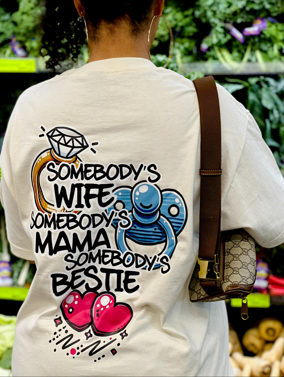 Back view of a woman wearing the white "Somebody's Wife, Mama, Bestie" Tee by Dass My Best Friend, shopping for vegetables in Whole Foods. The playful graphic on the back celebrates her roles as a wife, mom, and best friend, in a relaxed and stylish fit.