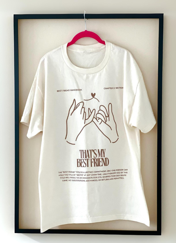 Full frontal view of the tan "That's My Best Friend" Tee by Dass My Best Friend, hanging inside a picture frame. The tee features a soft, relaxed fit with the bold "That's My Best Friend" design printed on the front, perfect for celebrating your bond with your best friend.