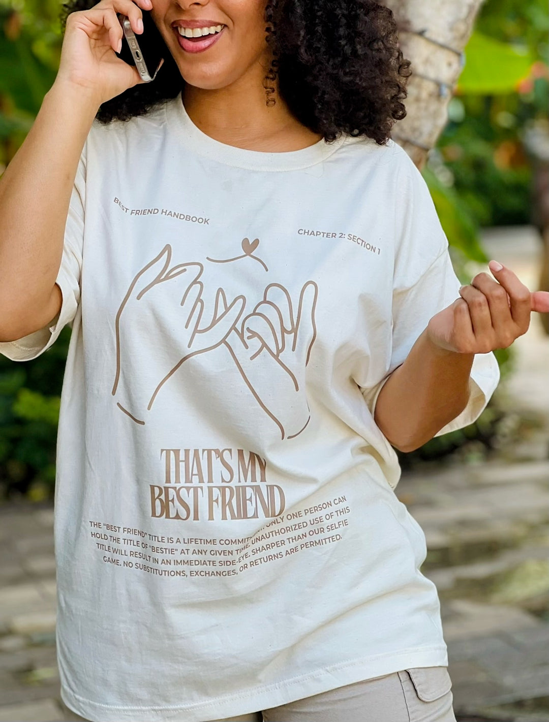 Woman talking on her cell phone wearing the tan "That's My Best Friend" Tee by Dass My Best Friend, frontal view. The tee features a soft, relaxed fit for all-day comfort, with the bold "That's My Best Friend" design printed on both the front and back.