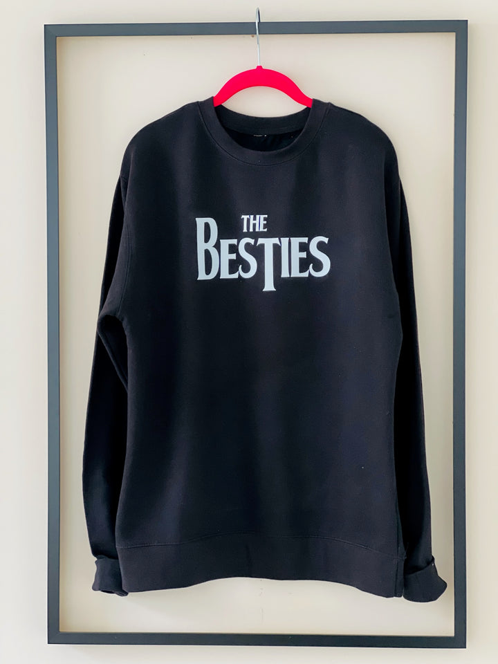 The Besties Crewneck in black by Dass My Best Friend, hanging inside a picture frame. This midweight sweater features a Beatles-inspired Besties design on the front. Made from an 80% cotton and 20% polyester blend, it's perfect for an oversized fit and a great gift for your bestie.