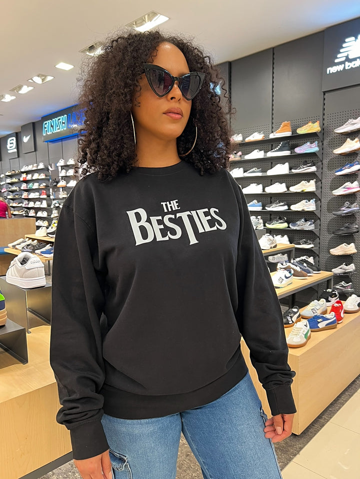 Woman wearing the black Besties Crewneck by Dass My Best Friend, standing in Footlocker. The Beatles-inspired Besties design is displayed on the front of the midweight sweater, combining comfort and style for a casual, everyday look.