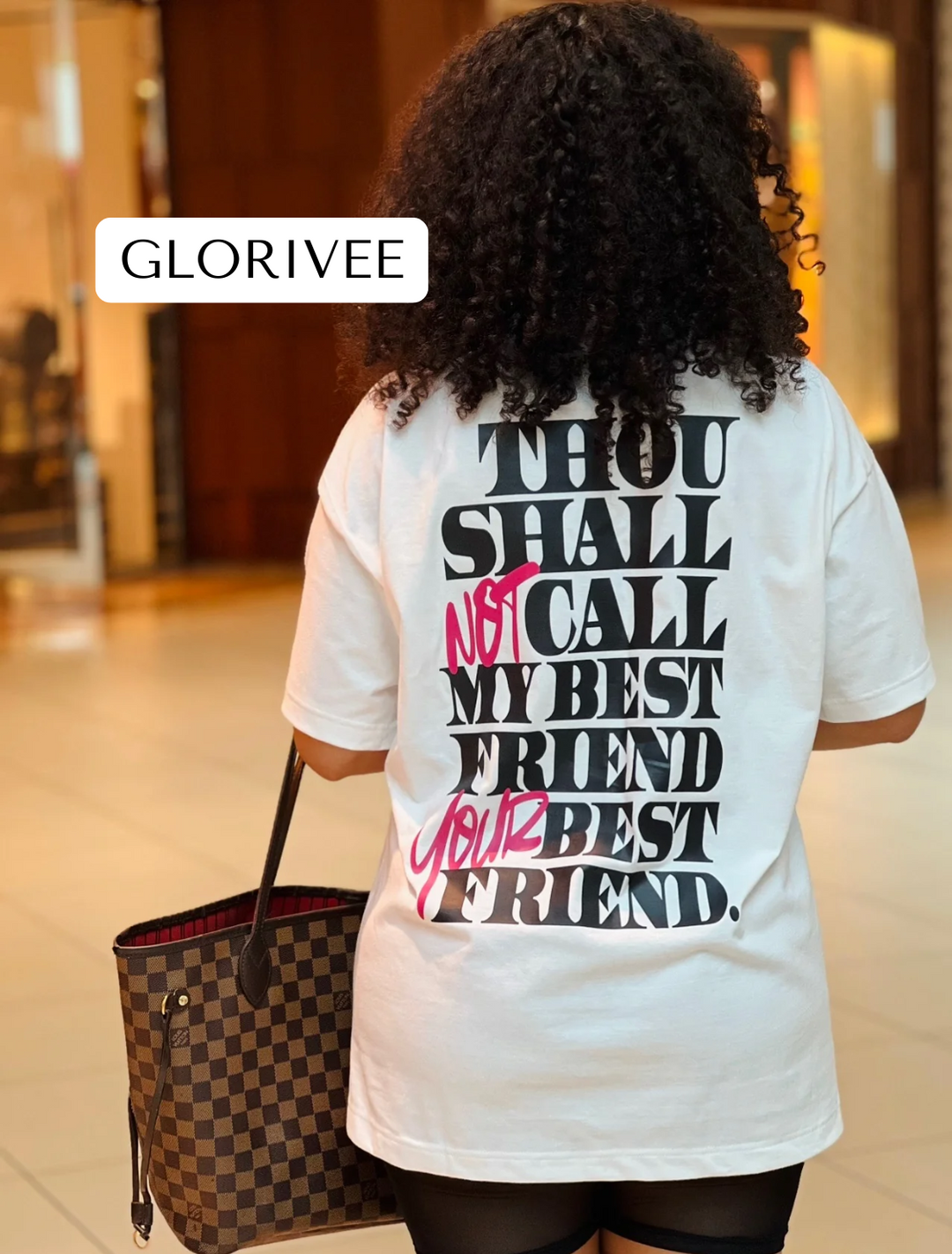 Glorivee walking in the mall holding a Louis Vuitton handbag, wearing the white "Thou Shall Not" Tee by Dass My Best Friend, rear view. The bold "Thou Shall Not" graphic is displayed on the back, highlighting the oversized fit and playful message.