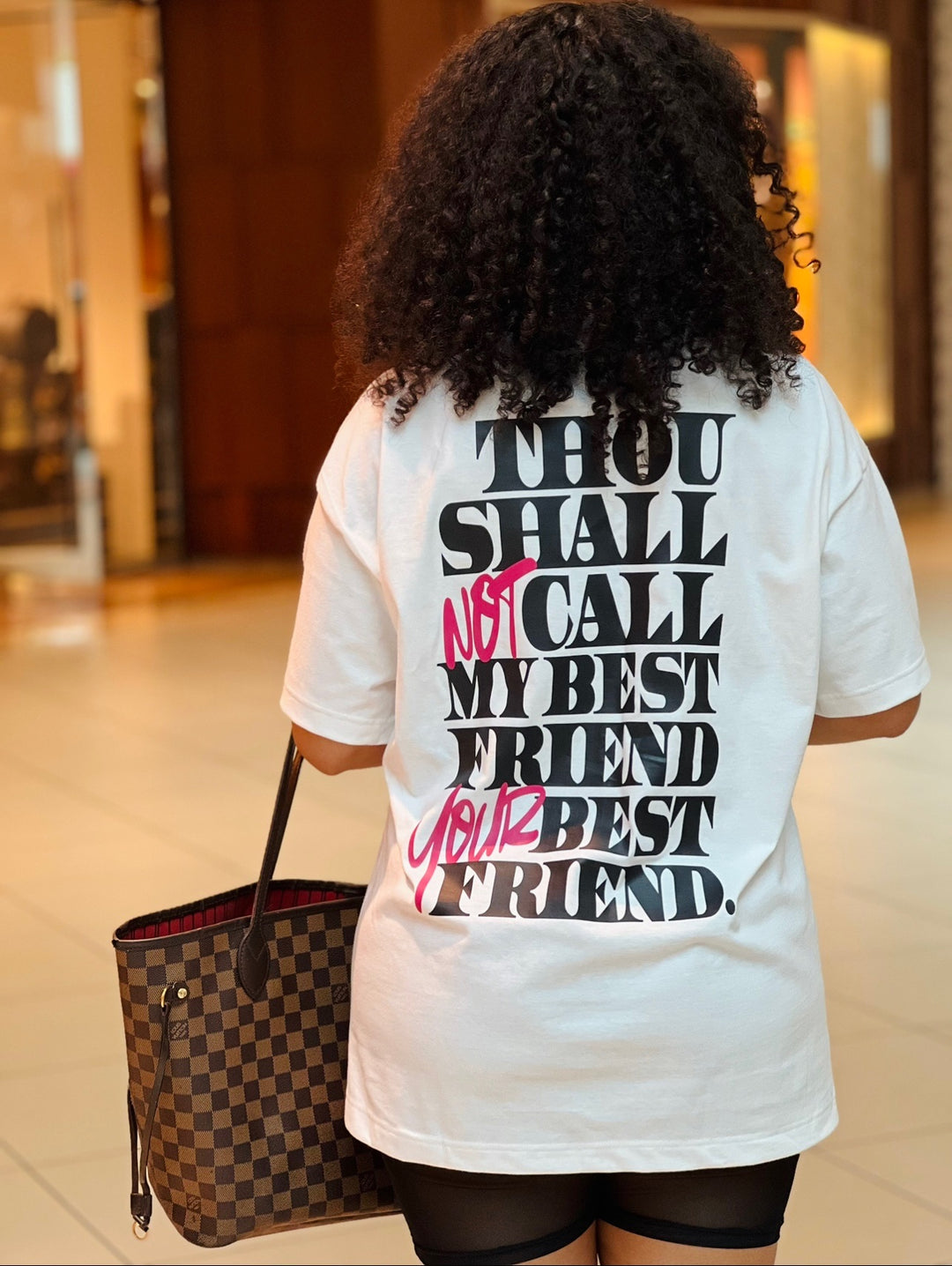 Woman walking in the mall holding a Louis Vuitton handbag, wearing the white "Thou Shall Not" Tee by Dass My Best Friend, rear view. The bold "Thou Shall Not" graphic is displayed on the back, highlighting the oversized fit and playful message.